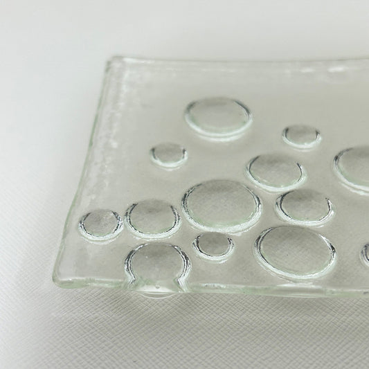 June Yoda「ガラス正方皿」
ガラス造形作家の June Yoda(依田淳子氏)による、水滴模様がほどこされた正方形の小皿作品。
小皿の脚が円形や楕円形のガラスで成形されており、上から眺めると、まるで輝く大粒の水滴がお皿を持ち上げているように見えます。